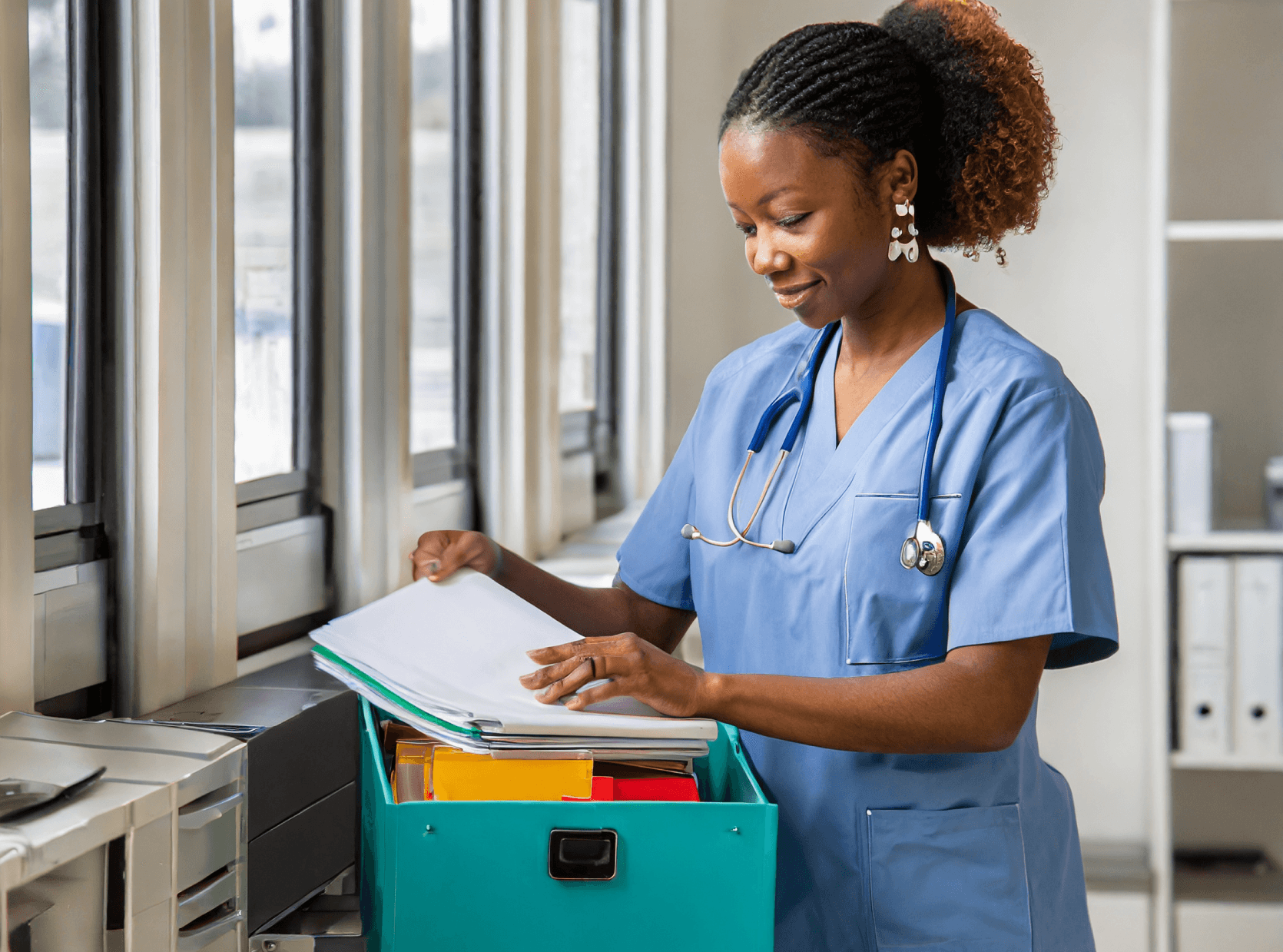 Healthcare worker in blue scrubs and stethoscope reviews files from a box in a well-lit room with large windows