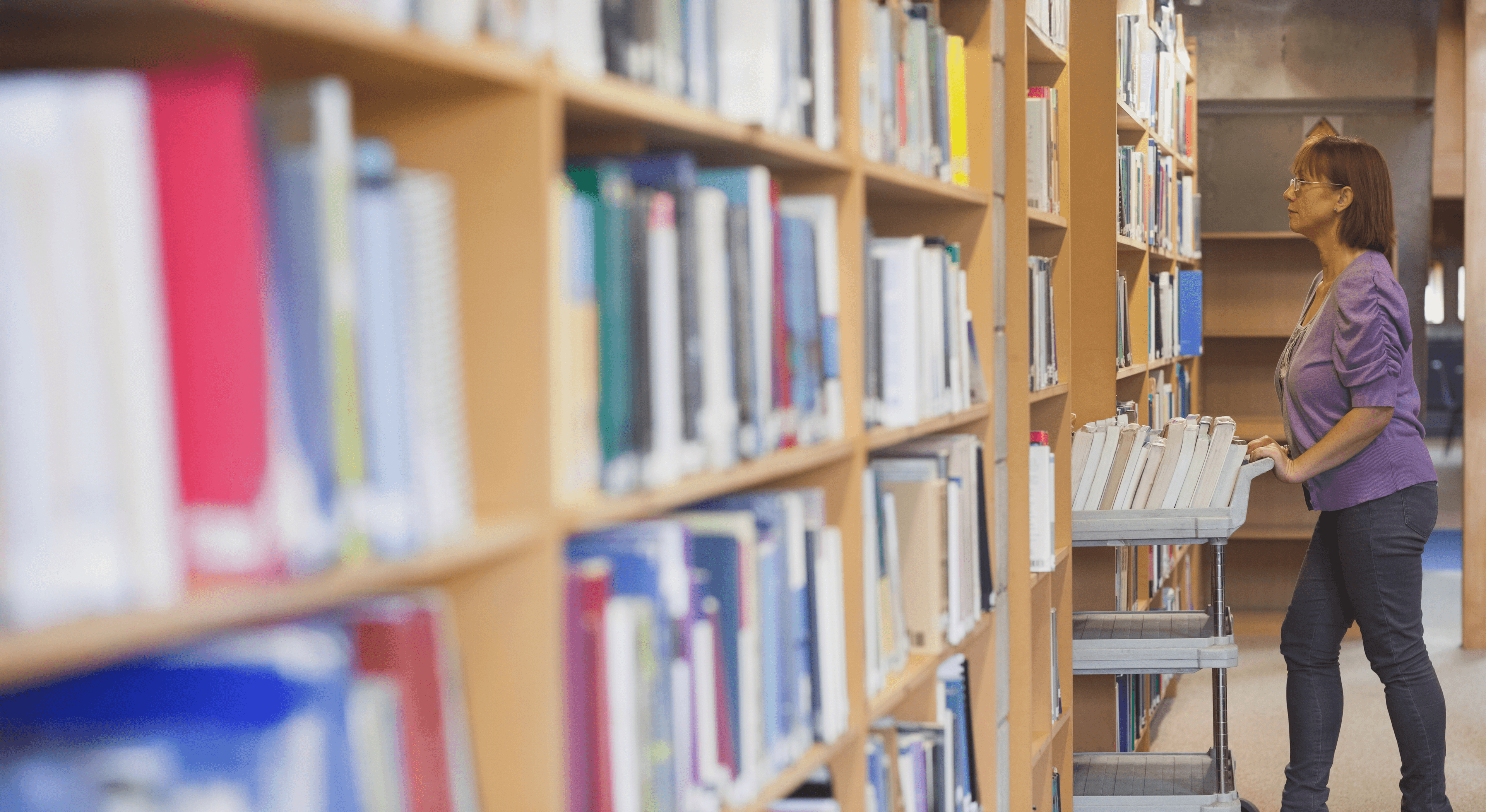 A librarian or archivist in a purple sweater and glasses organizing books on a cart in a library, surrounded by tall bookshelves filled with colorful books.